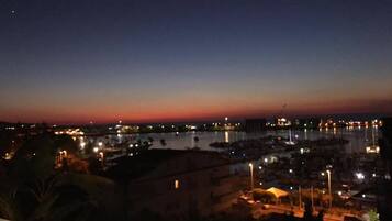 Vista nocturna de un puerto deportivo con barcos amarrados, una masa de agua tranquila y edificios iluminados a lo largo de la orilla.