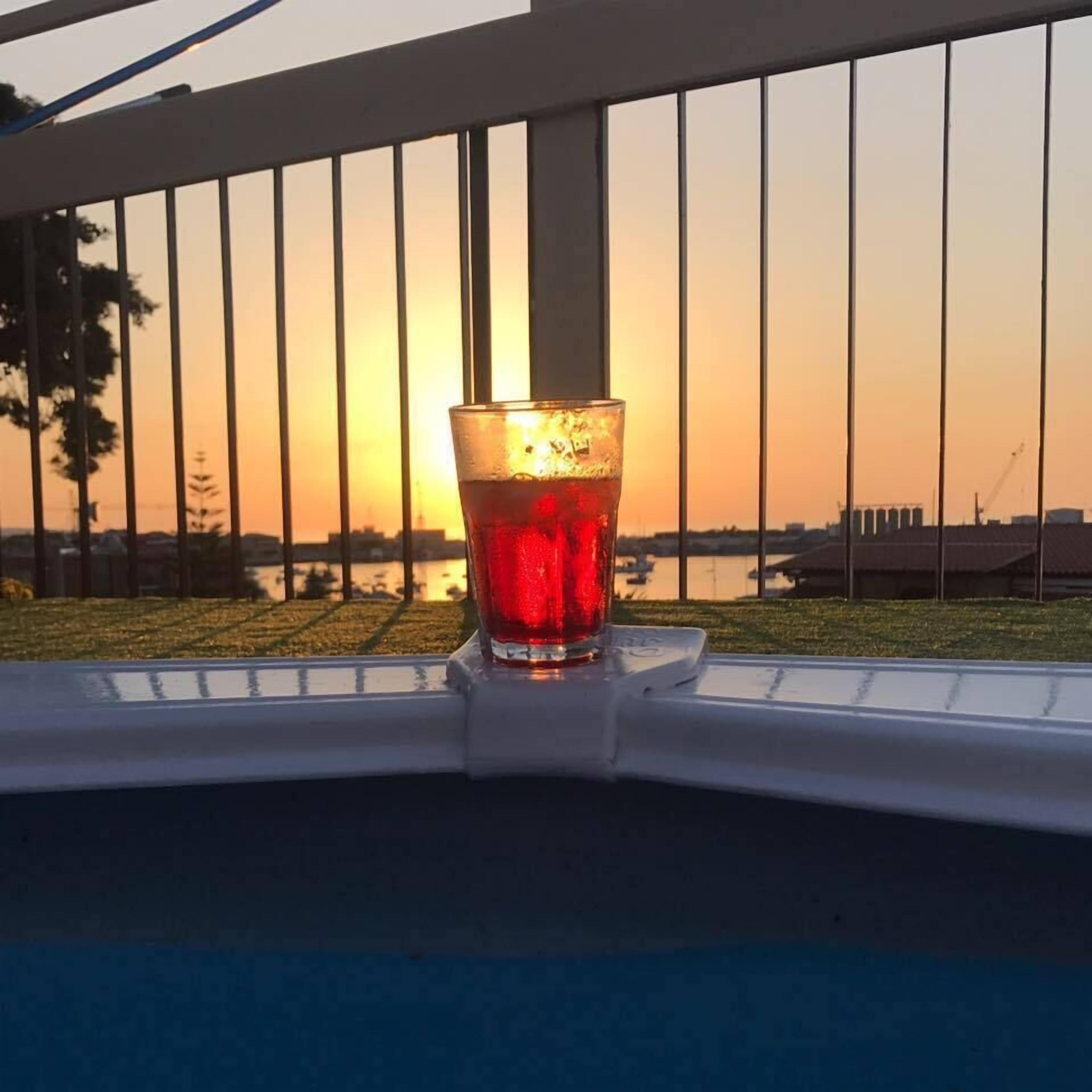 Ein Glas rotes Getränk auf einem weißen Tablett, mit Blick auf einen Sonnenuntergang durch ein Metallgitter.
