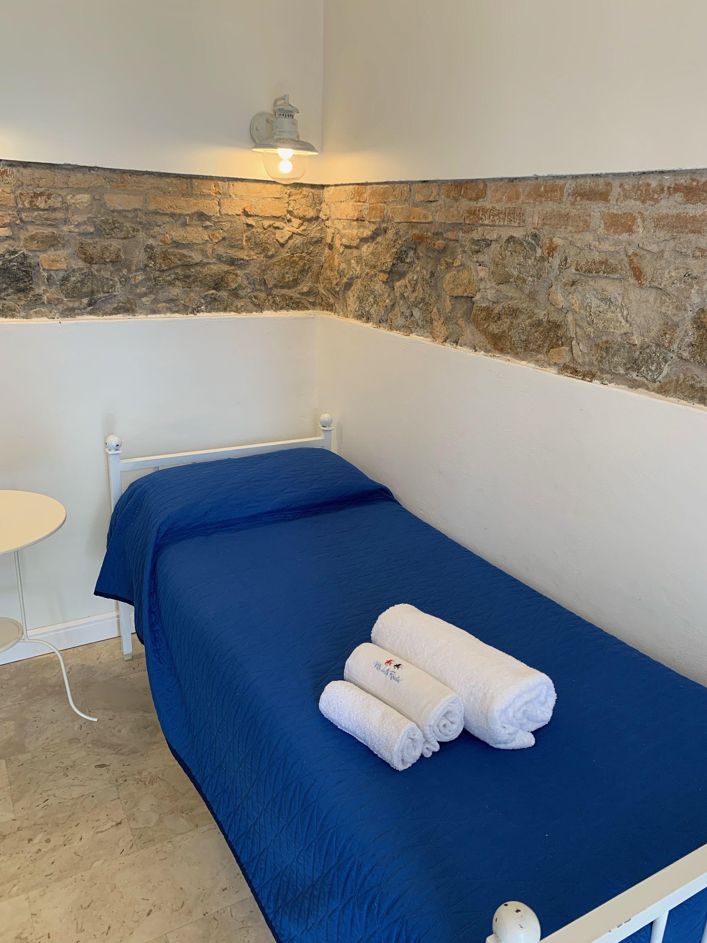 A single bed with a blue mattress and white bedding, a small round table, and a stone wall.