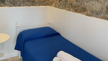 A single bed with a blue mattress and white bedding, a small round table, and a stone wall.
