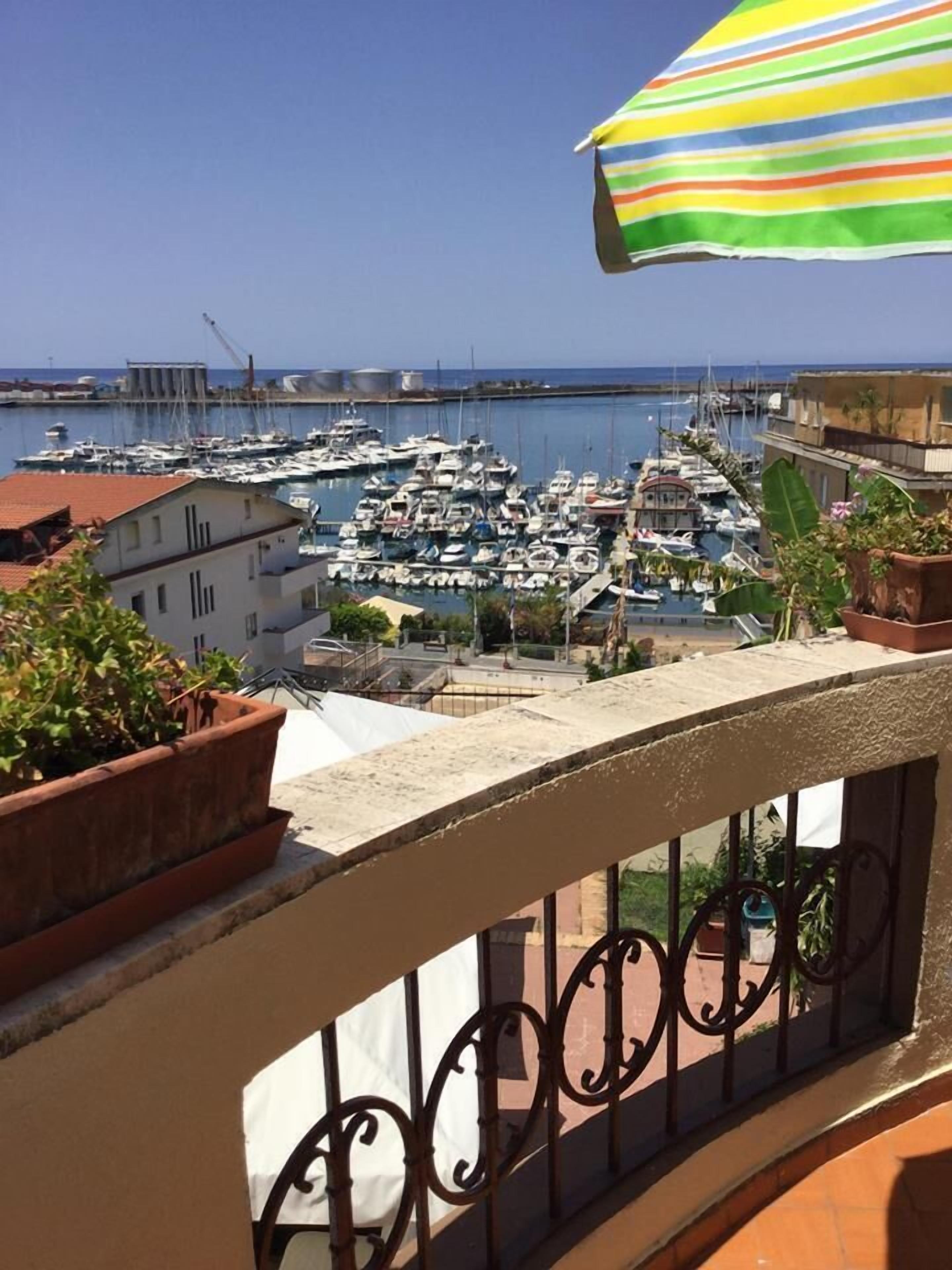 Un balcon donnant sur une marina où sont amarrés de nombreux bateaux, agrémenté de plantes en pot et surmonté d’un parasol rayé.