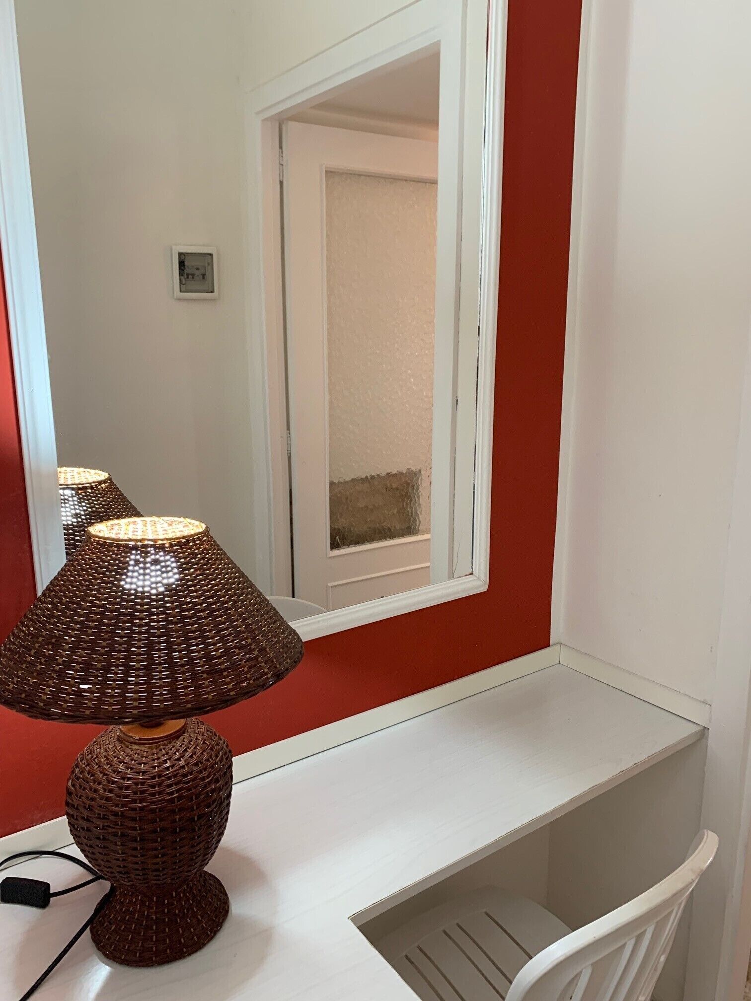 A corner desk with a woven lamp, a white chair, and a red wall.
