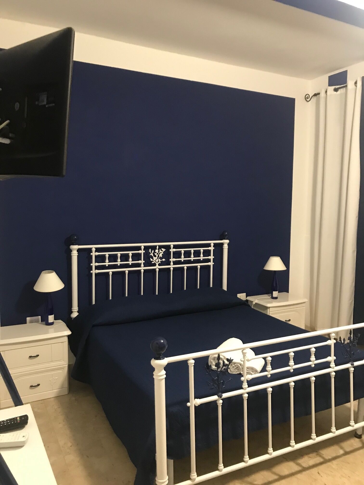 A bedroom with a white metal bed frame, a dark blue bedspread, a white bedside table with a lamp, and a white nightstand with a lamp.