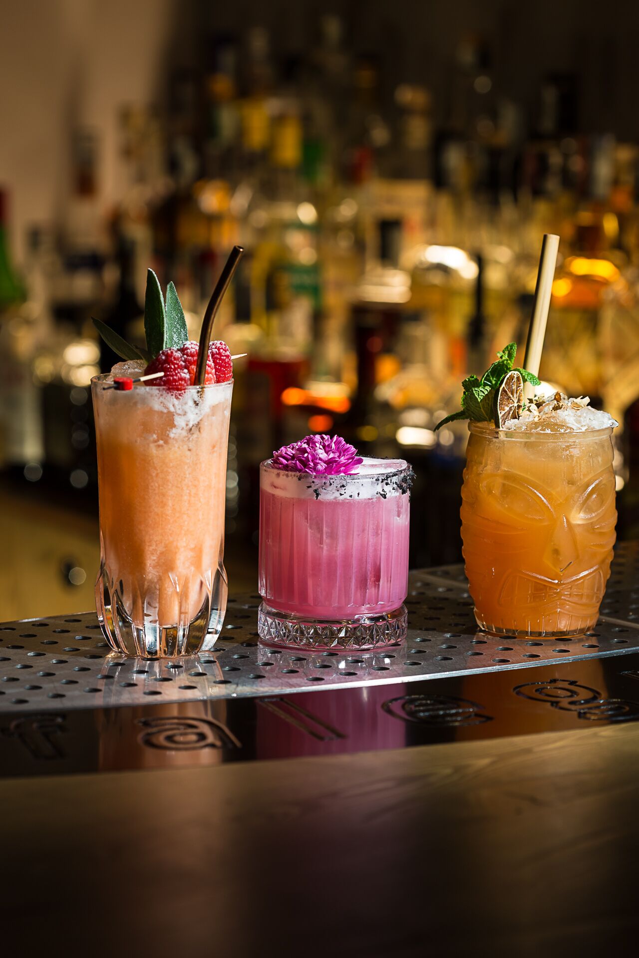 Drei Cocktails auf einer Bar, im Hintergrund verschwommene Flaschen.