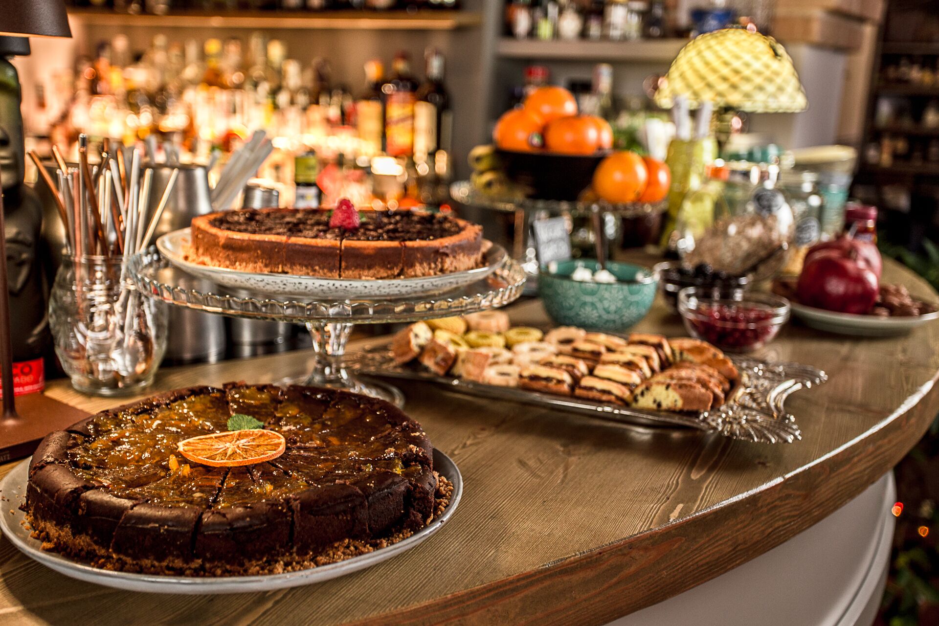 Una zona de bar con diversos postres a la vista, como pasteles y repostería, y una selección de botellas y vasos al fondo.