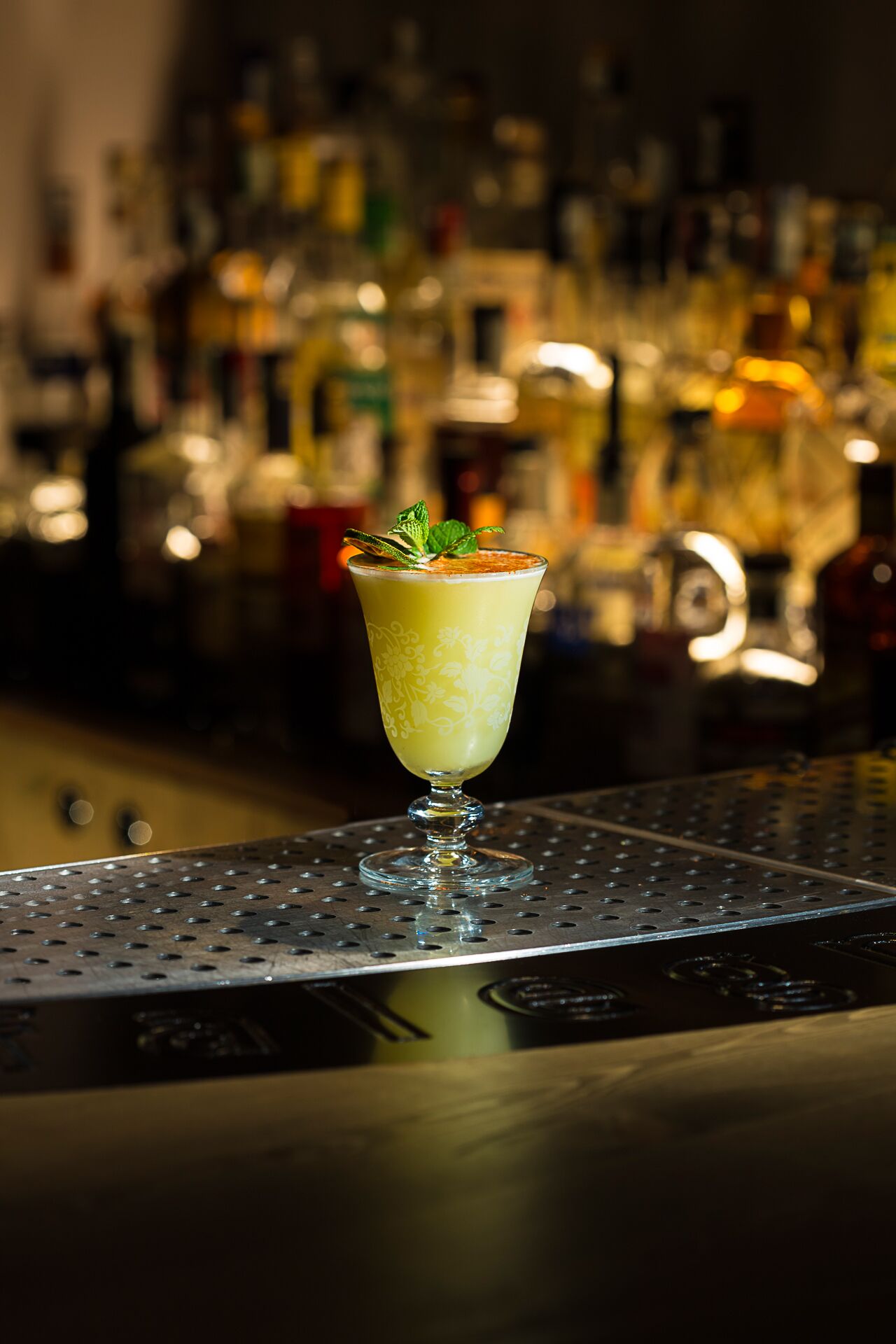 A cocktail in a glass with a decorative pattern, garnished with mint leaves, on a bar counter with a blurred background of various bottles.