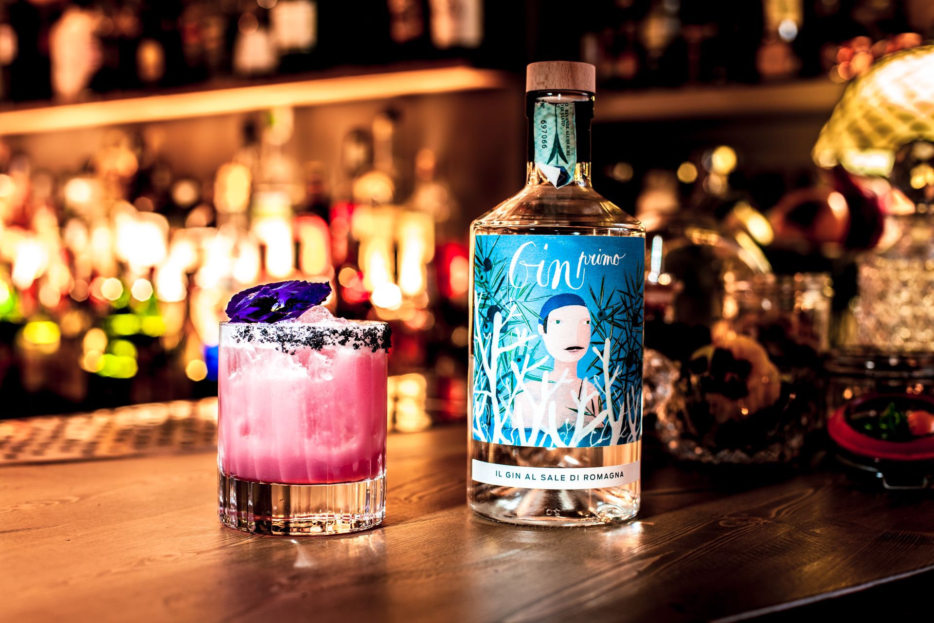 A bottle of gin and a pink cocktail on a bar counter with a blurred background of other bottles.