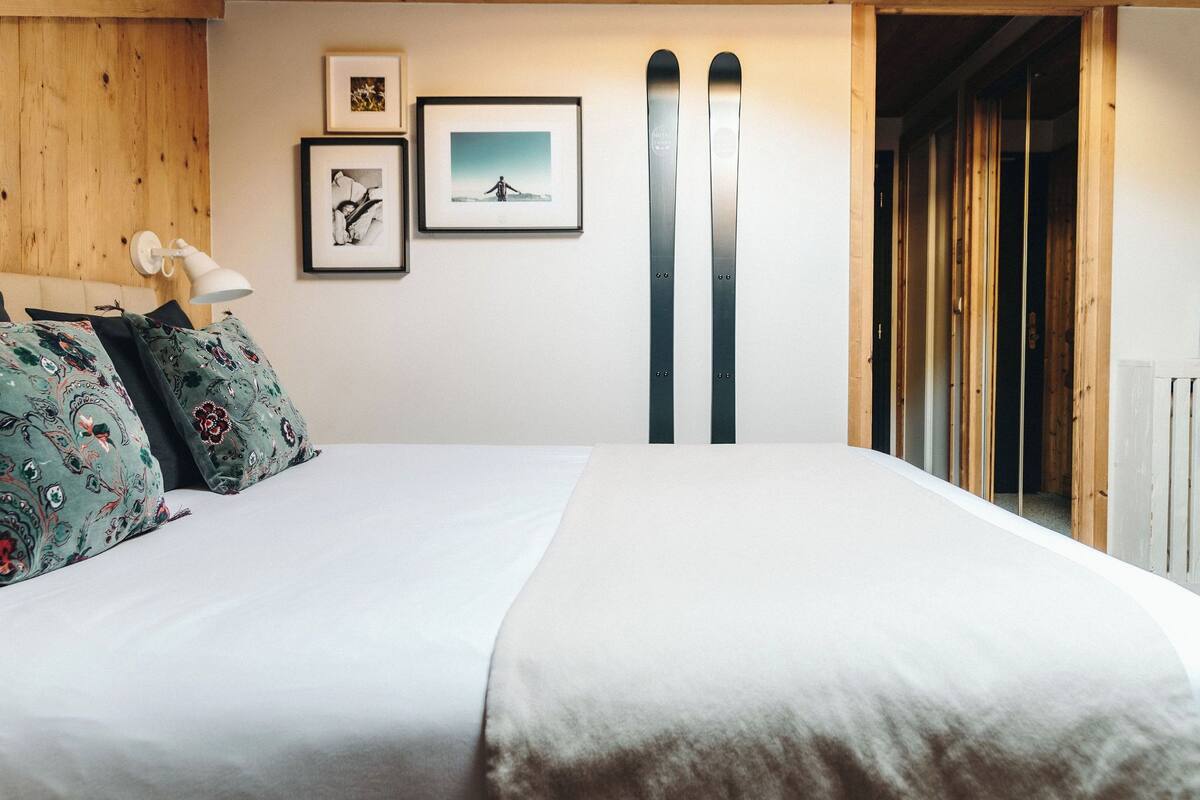 Une chambre d’hôtel avec une tête de lit en bois, deux tableaux encadrés au mur et une paire de skis appuyés contre le mur.