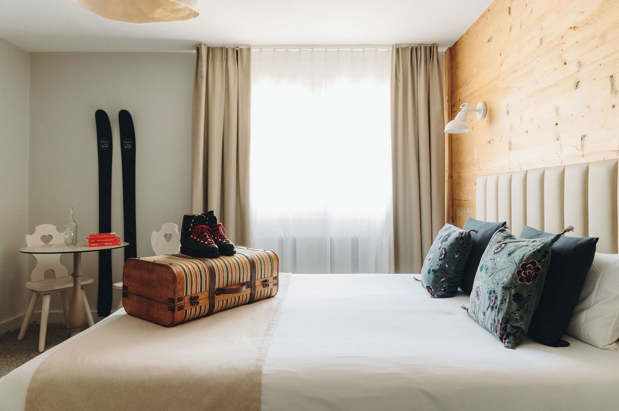 A hotel room with a bed, a suitcase, skis, a table, and a chair.