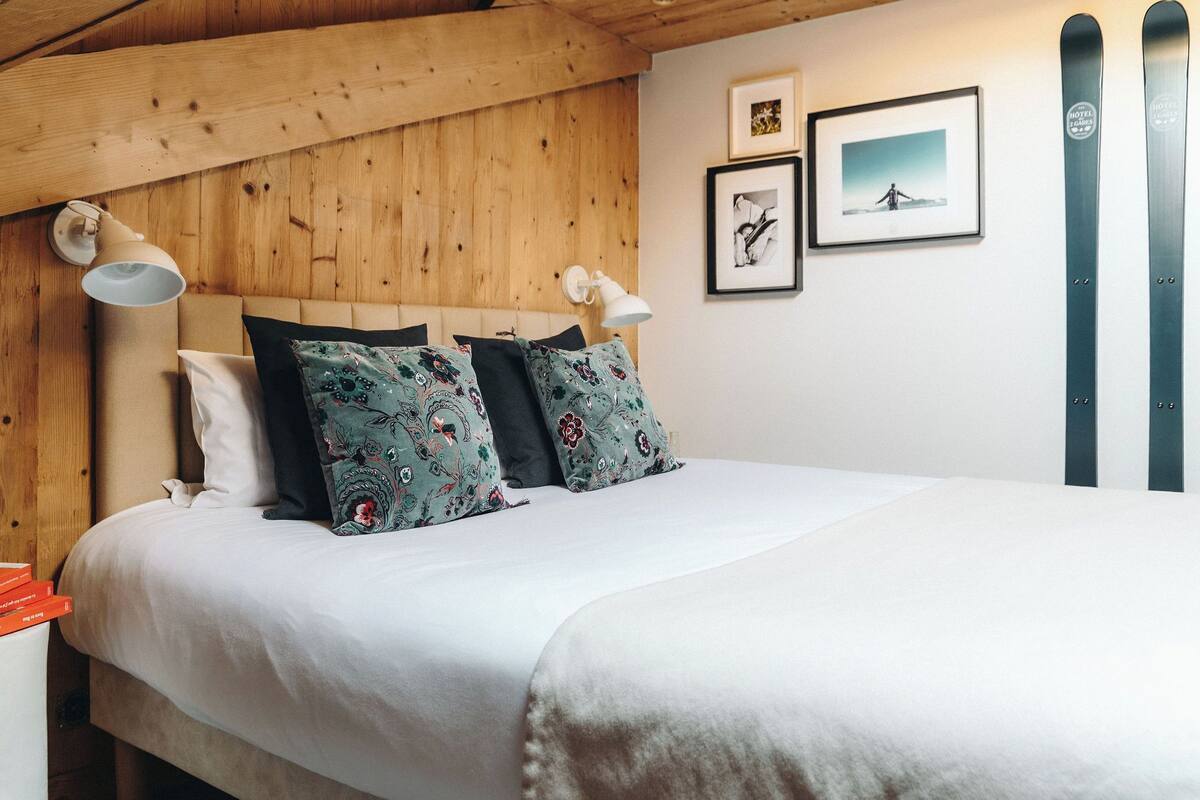 Une chambre avec un plafond en bois, un lit recouvert de linge de lit blanc, deux coussins à motifs et deux lampes fixées au mur.