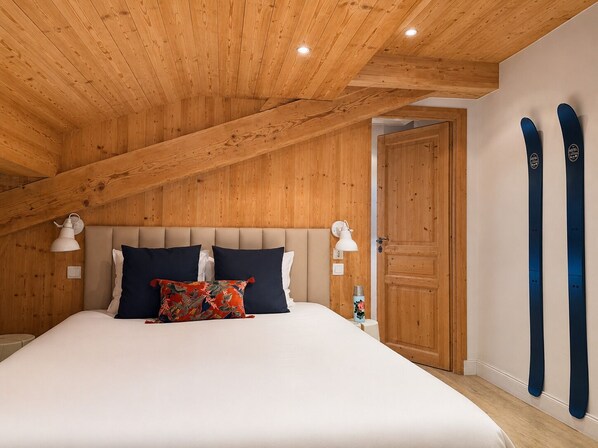 A bedroom with a wooden ceiling, a bed with white bedding, two wall-mounted lamps, and a door with skis leaning against it.