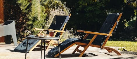 Dos sillones de descanso con respaldos de lona y armazones de madera, una mesita con una taza y un plato, y una silla blanca en un entorno exterior soleado.