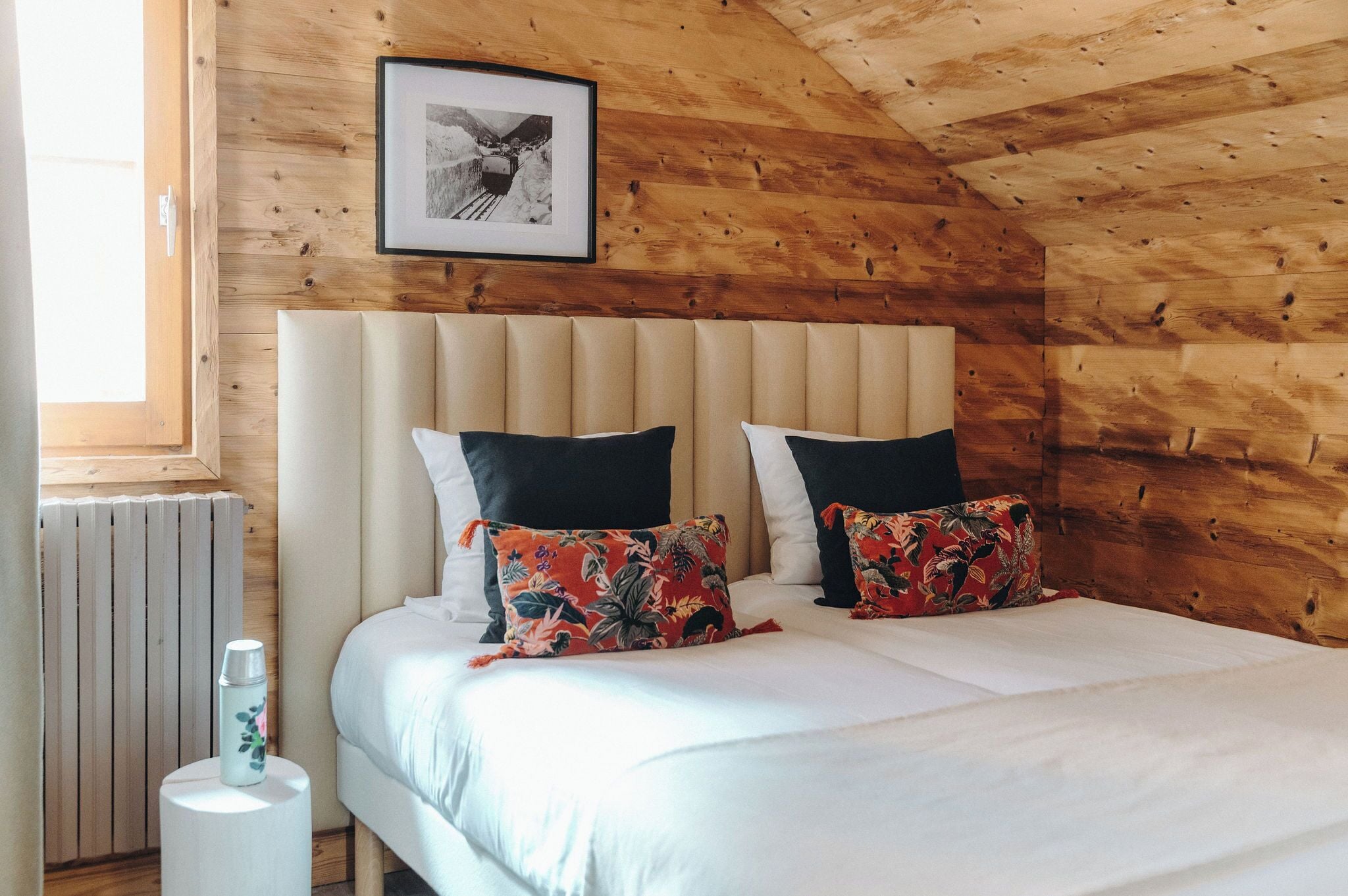 A wooden-paneled room with a bed featuring a white mattress, two black pillows, and a floral pillow. A framed picture hangs on the wall above the bed.