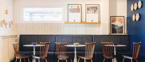 A restaurant interior with a dark-colored booth seating area, wooden chairs, and a small round table with a bowl.