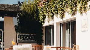 Un área de estar al aire libre con sillas de mimbre y un otomano blanco y mullido, un ventanal grande y una pared decorada con plantas colgantes.