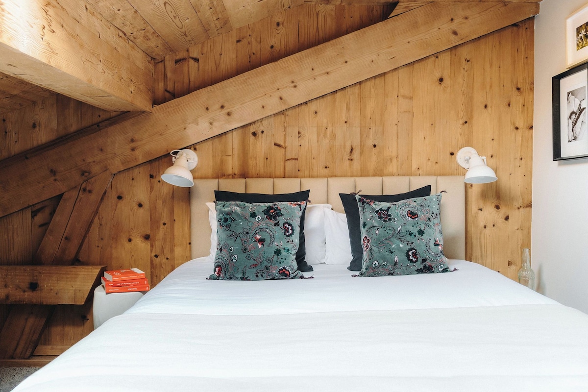 A bedroom with a wooden ceiling, a bed with white bedding, two patterned pillows, and bedside lamps.