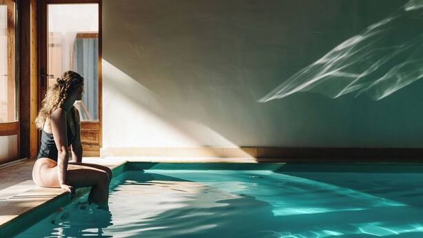 Una persona sentada junto a una piscina, con sombras proyectadas por la luz del sol en la pared.
