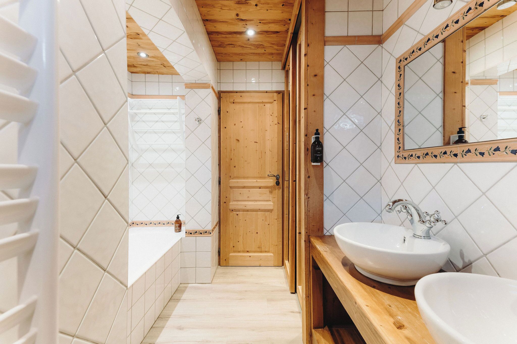 A bathroom with a wooden door, white tiled walls, a large mirror, and two sinks.