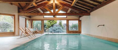 An indoor swimming pool with wooden beams, large windows, and a view of trees.