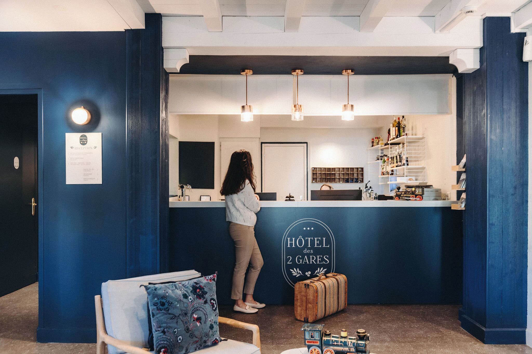 Una recepción con un cartel que dice "HOTEL des 2 GARES" y una mujer de pie detrás. Hay estantes con botellas y una maleta en el suelo.