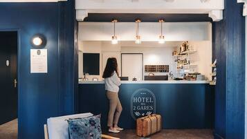 Una recepción con un cartel que dice "HOTEL des 2 GARES" y una mujer de pie detrás. Hay estantes con botellas y una maleta en el suelo.