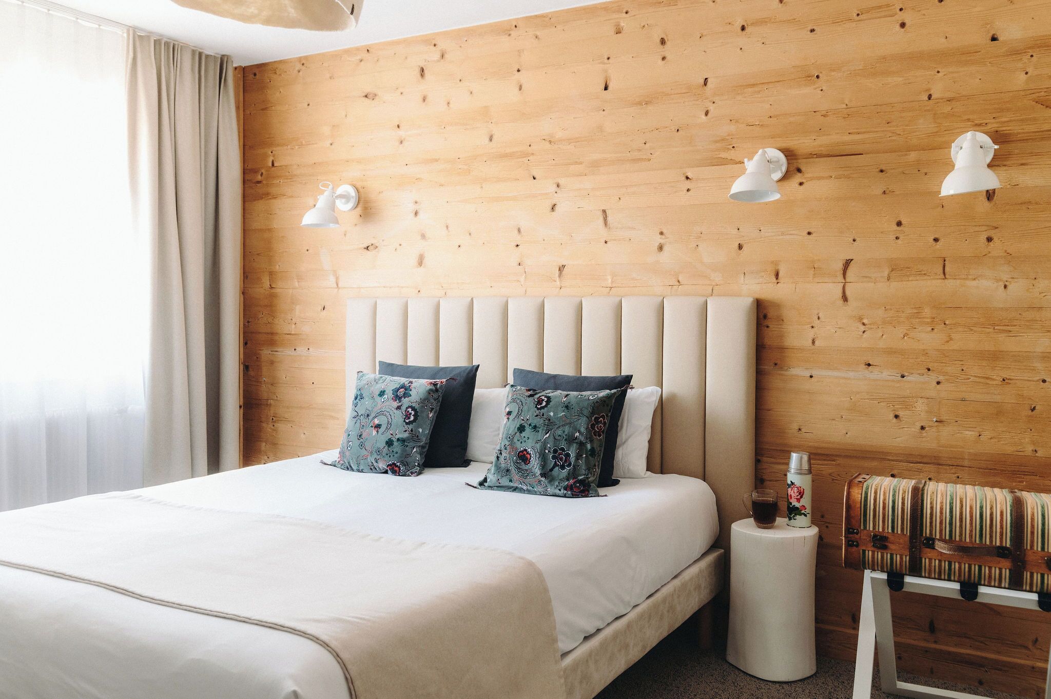 Habitación de hotel con pared de madera, una cama grande con dos almohadas, una mesita de noche con un termo y una silla pequeña.