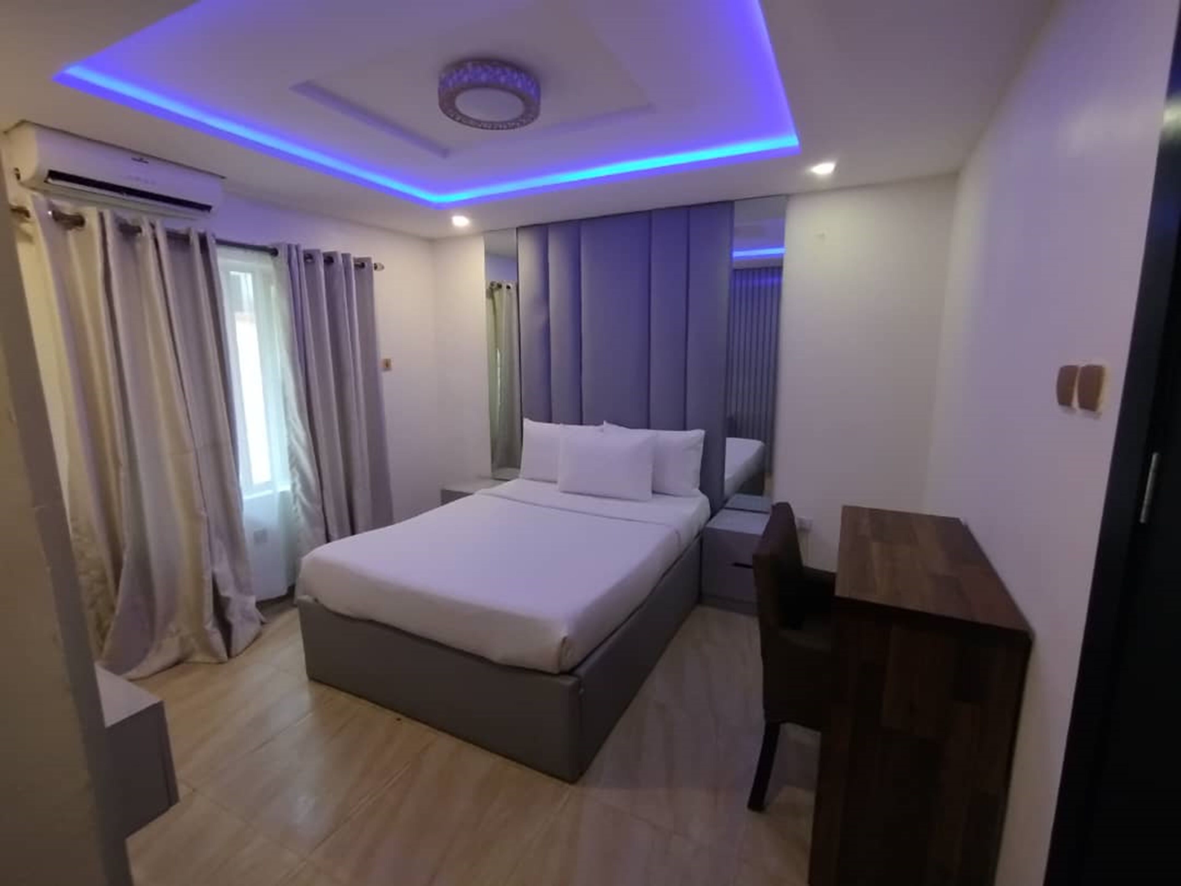 A modern hotel room with a large bed, a desk, and a ceiling with blue LED lighting.