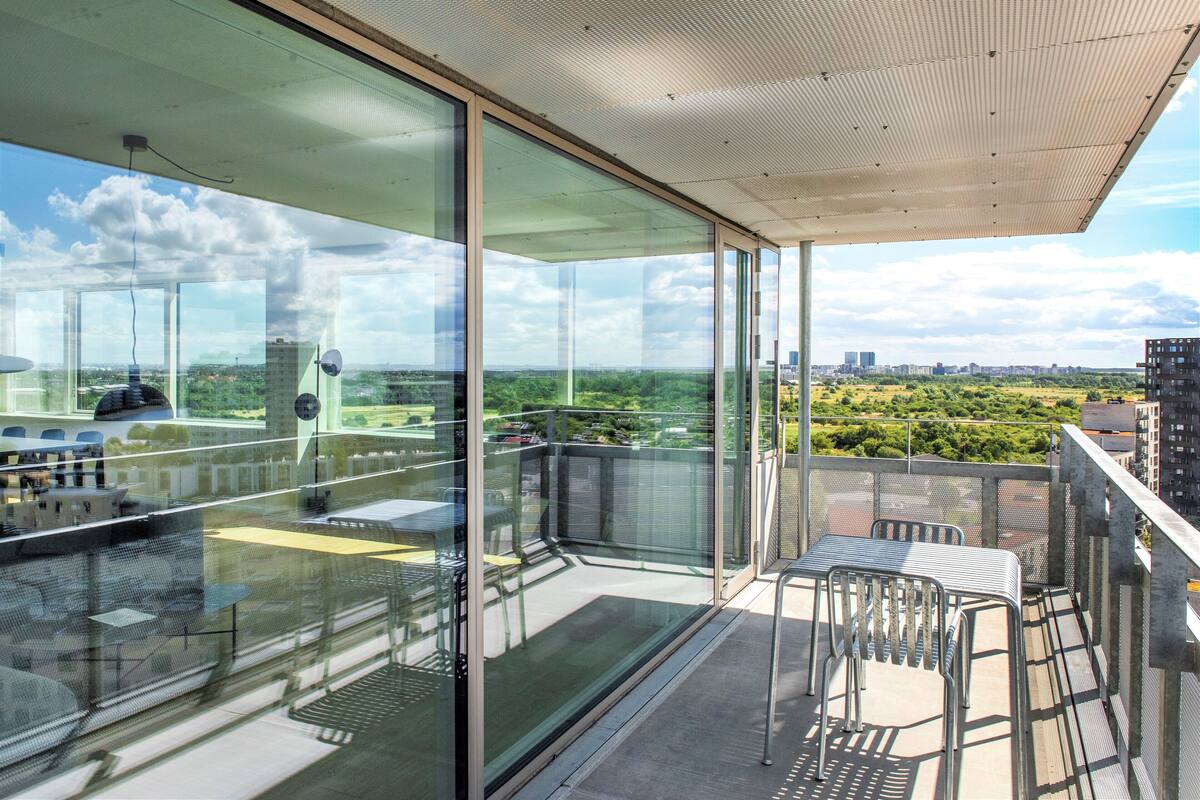 Un balcón moderno con paredes de vidrio, que ofrece vistas a un paisaje urbano y zonas verdes.