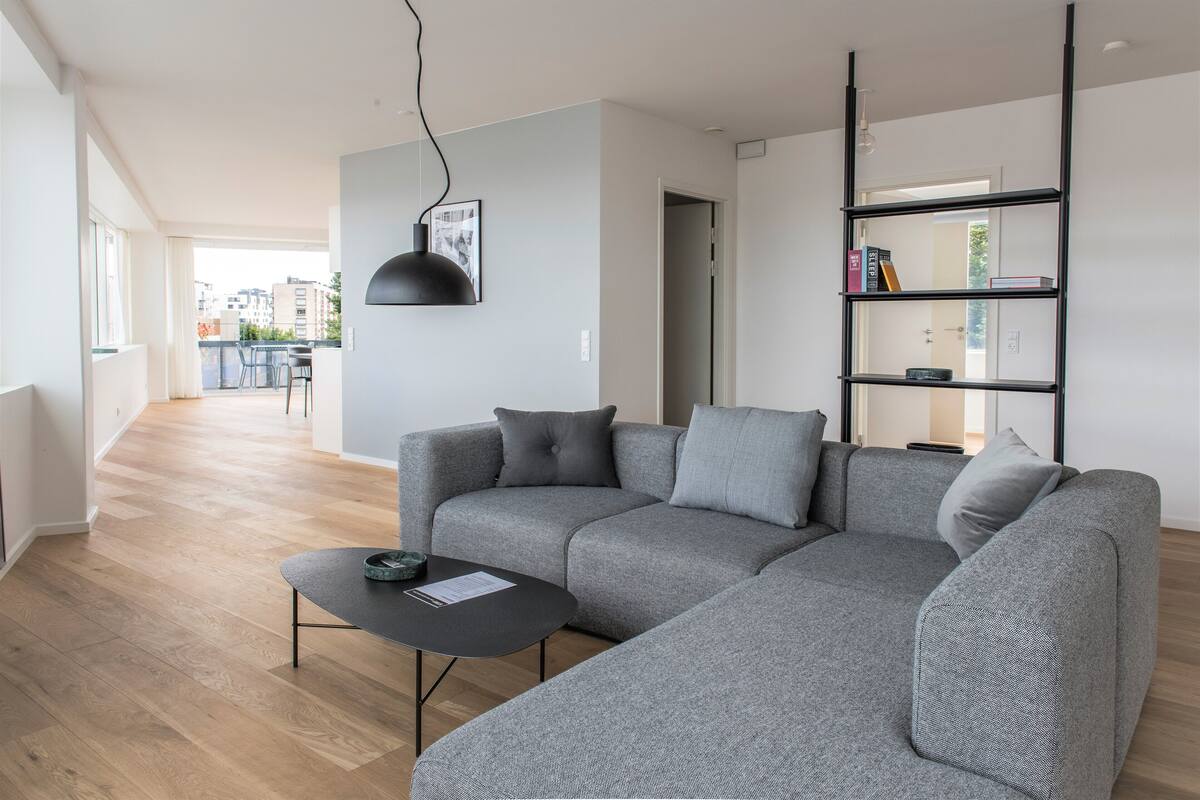 Una sala de estar moderna con un sofá seccional gris, una mesa de centro negra y una lámpara colgante negra. Hay una estantería con libros y objetos decorativos. El piso es de madera y hay vistas al exterior a través de ventanas grandes.