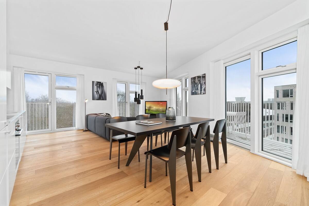 Un comedor amplio con suelo de madera, una mesa de comedor oscura y sillas modernas. Hay ventanas grandes que ofrecen vistas a la ciudad.