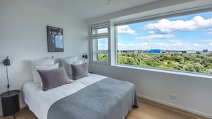Un dormitorio con un ventanal grande, una cama con ropa de cama blanca y gris, una mesita de noche y una lámpara de pared.