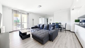 Een moderne woonkamer met een grijze hoekbank, een eettafel met stoelen, en een keuken met een koffieapparaat.