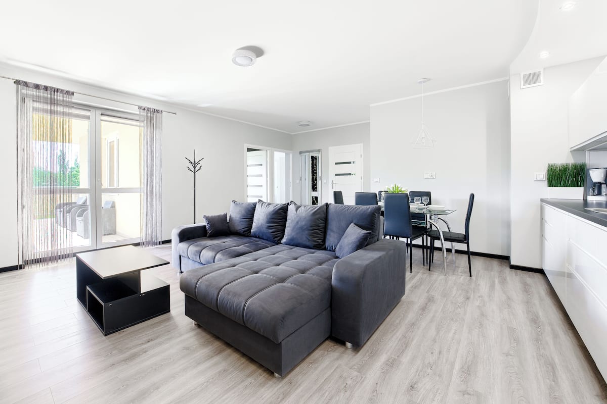 Een moderne woonkamer met een grijze hoekbank, een eettafel met stoelen, en een keuken met een koffieapparaat.
