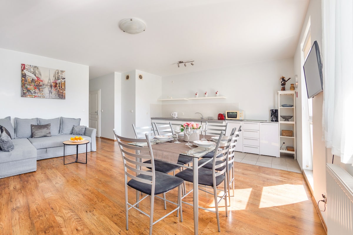 Een moderne woonkamer met een grijze bank, een eettafel met stoelen, een keuken met witte kastjes en een schilderij van een stadsgezicht.