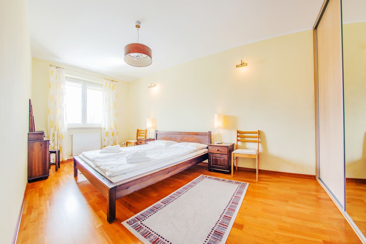 Een slaapkamer met een houten bed, nachtkastjes met lampen, een stoel, een radiator en een raam met gordijnen.