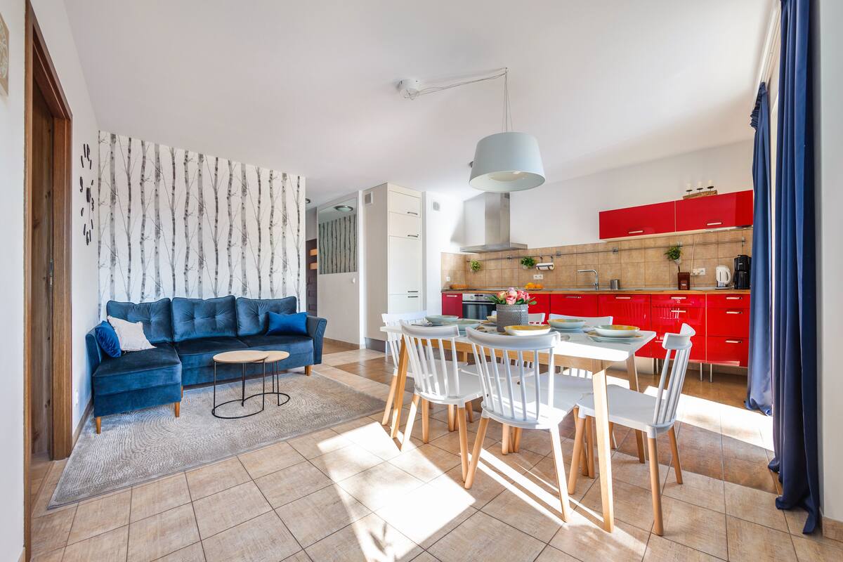 Een moderne woonkamer met een blauwe bank, een eettafel met stoelen, en een keuken met rode kasten.