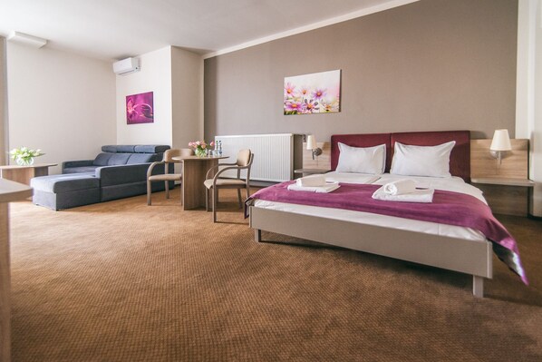 Habitación de hotel con una cama grande, un sofá, una pequeña mesa con sillas, un radiador y obras de arte en las paredes.