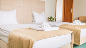 Habitación de hotel con dos camas, cada una con colcha color beige y ropa blanca. Se observa una puerta de madera y una mesita con una toalla doblada.