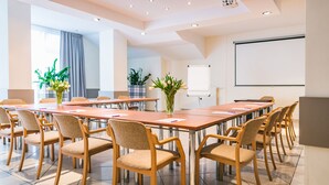 Un salón de conferencias con varias mesas y sillas, una pantalla para proyector y plantas en macetas.