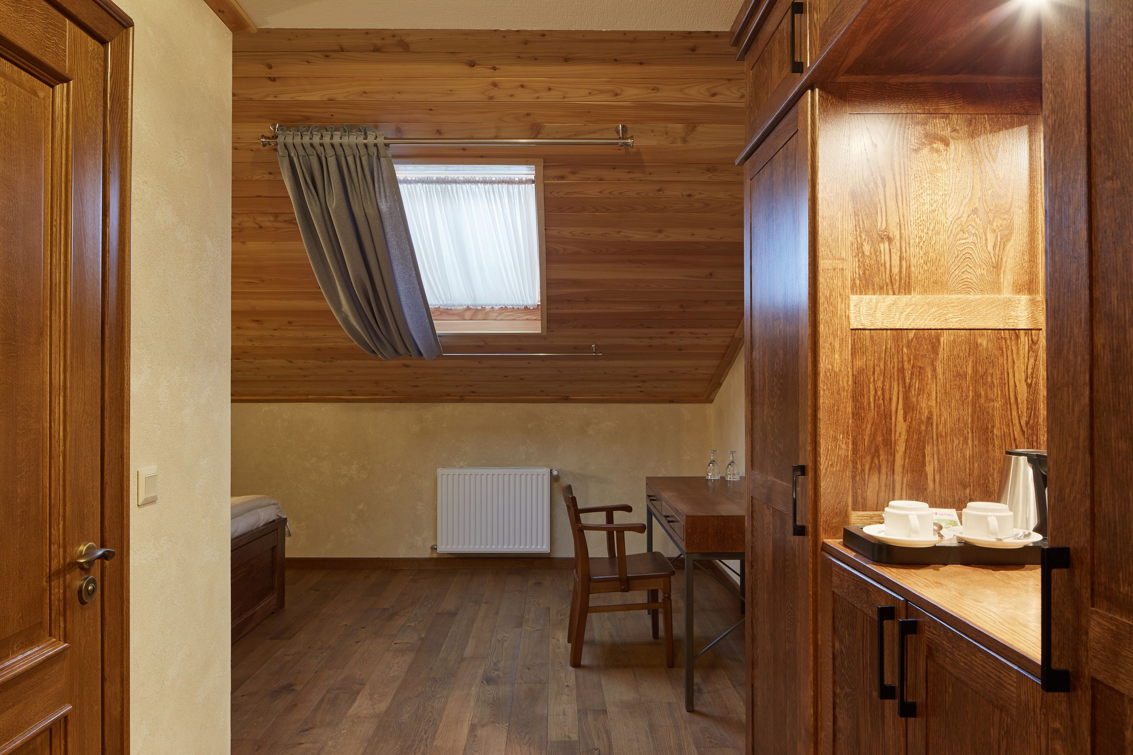 Una habitación con paneles de madera, techo inclinado, ventana con cortinas, radiador, una silla de madera, una mesita con tazas y un hervidor, y un armario de madera.