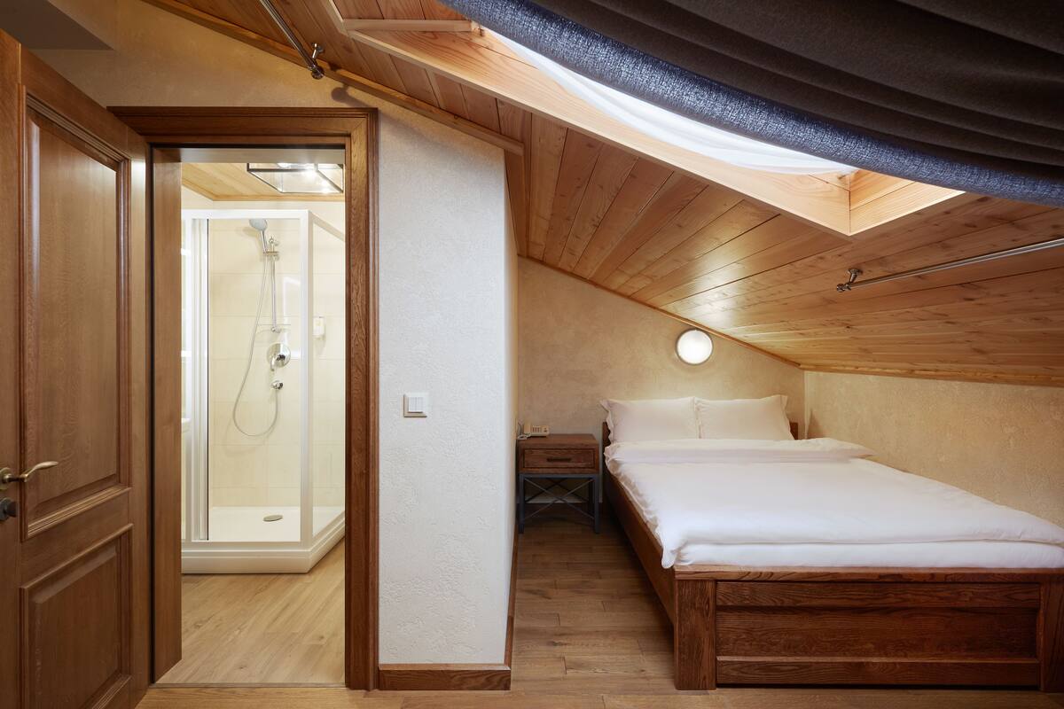 A bedroom with a wooden ceiling, a bed, a bedside table, a nightstand, and a walk-in shower.