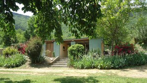 A small, single-story building with a sloped roof, surrounded by lush greenery and flowering plants. - Camping Le Roc Qui Parle (Nant)