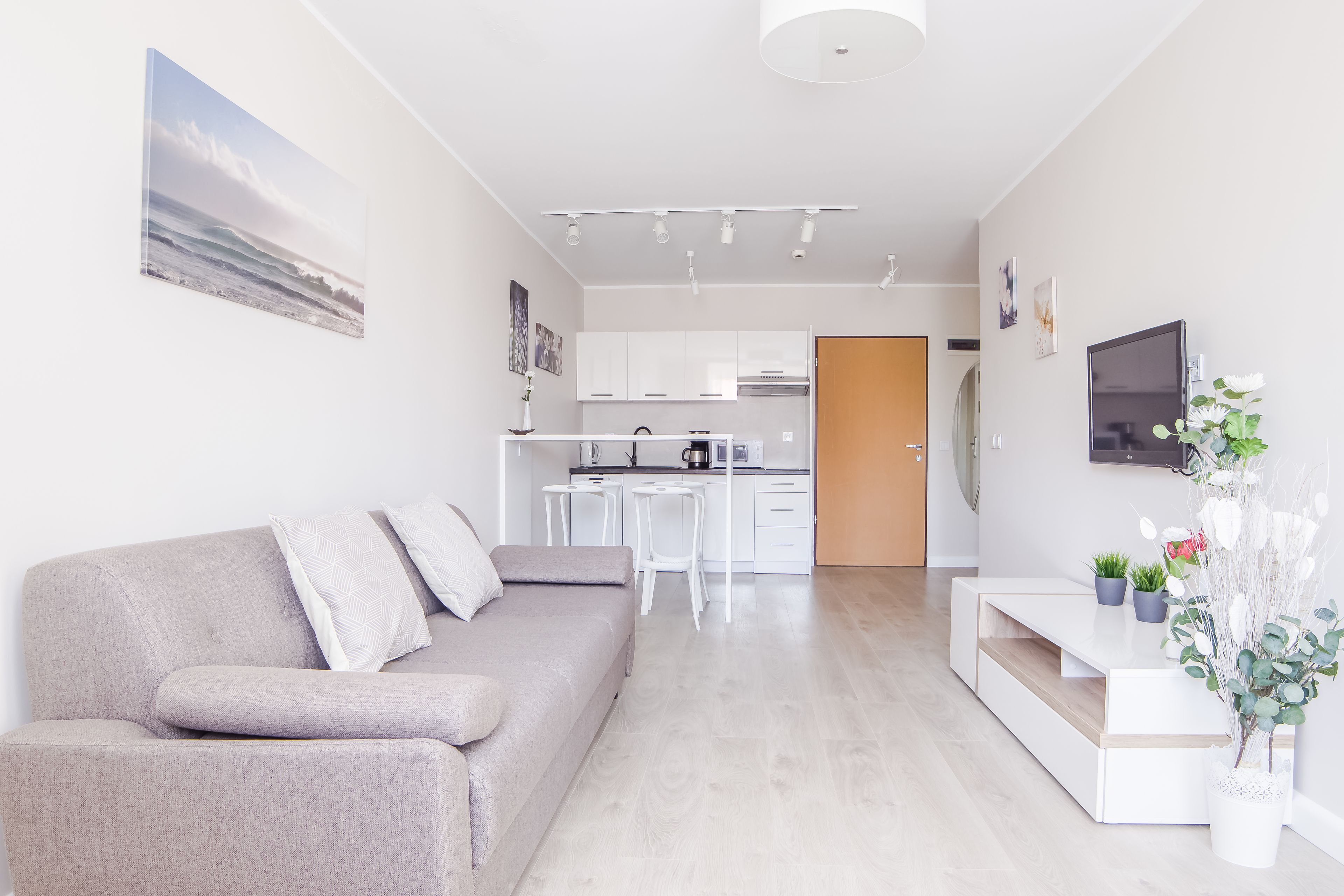 Una sala de estar moderna con un sofá gris, una mesa de comedor con sillas, una cocina al fondo y un televisor montado en la pared.