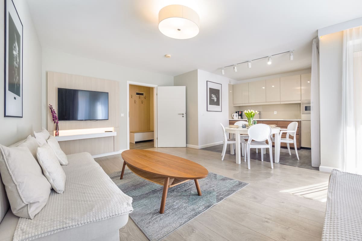 Una sala de estar moderna con sofá, una mesa de centro y un televisor montado en la pared. Hay una zona de comedor con mesa y sillas, y una cocina al fondo.