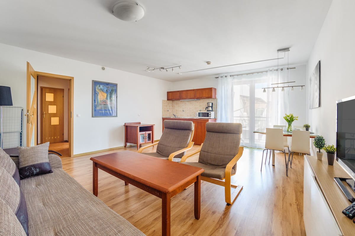 Una sala de estar moderna con un sofá, un sillón, una mesa de centro y un televisor. Se observa un área de cocina con armarios y un microondas.