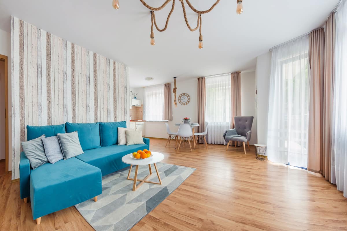 Una sala moderna con un sofá azul, una mesa redonda de centro con naranjas, una araña y una zona de comedor con una mesa y sillas blancas.