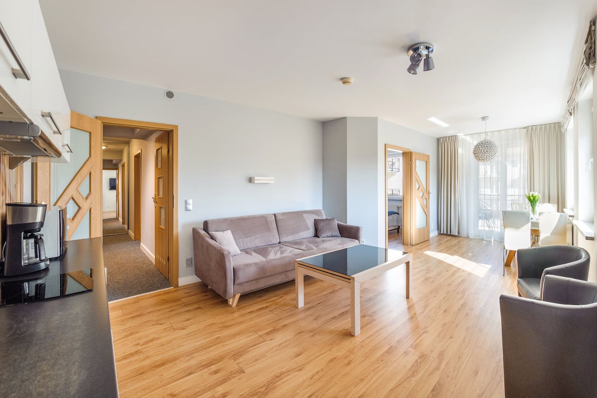 Una sala de estar moderna con sofá, mesa de centro y sillas. Incluye un área de cocina con cafetera y una mesa de comedor con sillas. El ambiente tiene piso de madera y una escalera que lleva a otro nivel.
