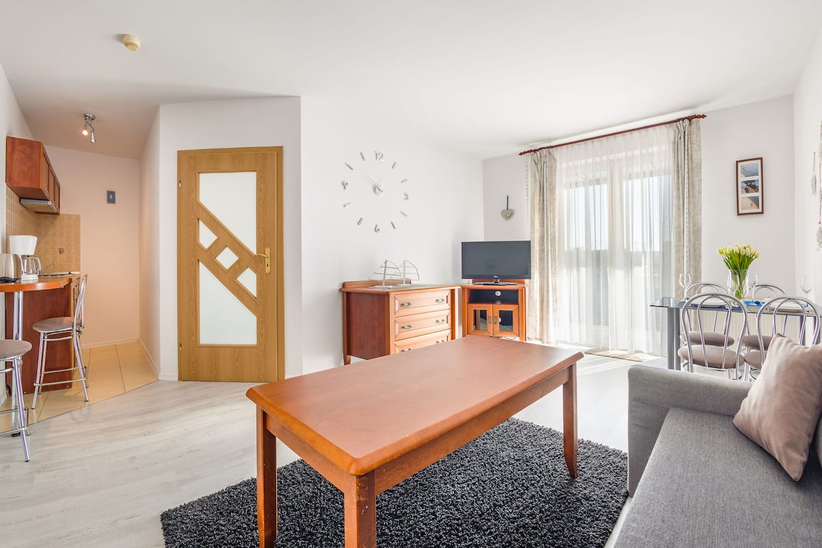 Una sala de estar moderna con una mesa de centro de madera, un sofá gris y un televisor. Hay una zona de cocina con una cafetera y sillas.
