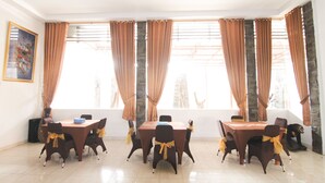 A dining area with a table set for six, surrounded by chairs with yellow sashes, large windows with curtains, and a framed picture on the wall. - Grand Ciwareng Inn (Purwakarta)