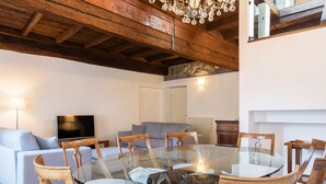 Uma sala de estar moderna com uma mesa de jantar de vidro rodeada por cadeiras de madeira, um sofá cinzento e uma televisão.